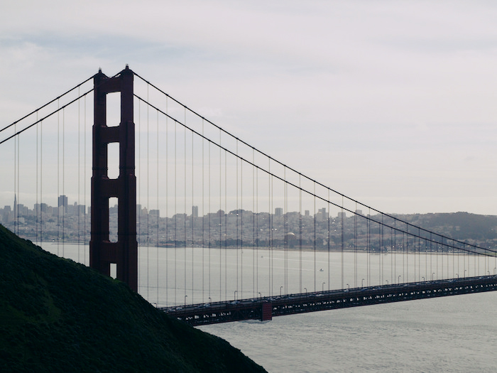A view of the Golden Gate Bridge from a bluff facing the city; San Francisco's high-rises are visible between the bridge beams on a slightly foggy day.